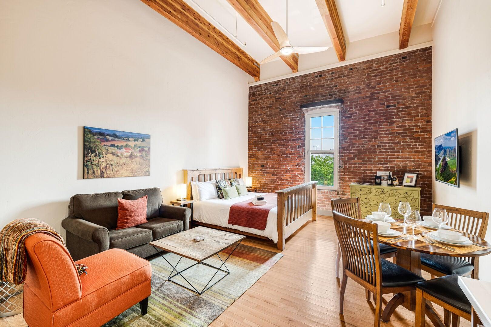 Modern studio apartment featuring exposed wooden ceiling beams and a brick accent wall with a tall window.