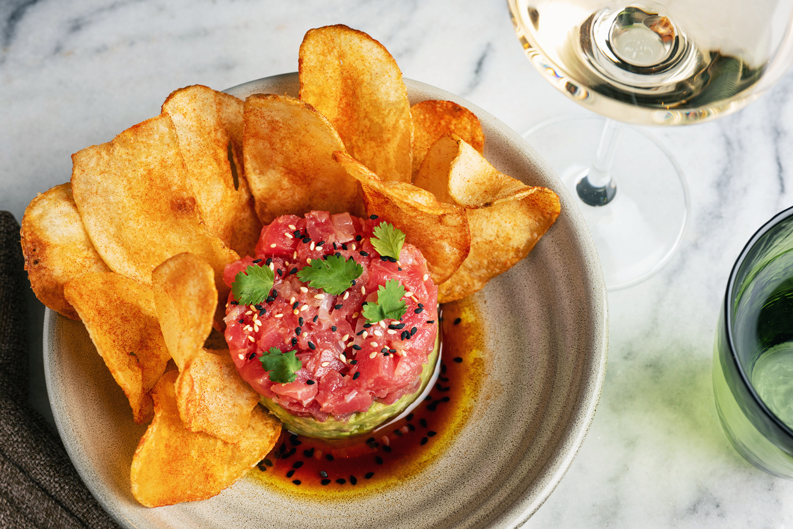 Plated appetizer featuring a round serving of tuna tartare garnished with cilantro and sesame seeds, surrounded by golden potato chips next to a stemmed glass of white wine.