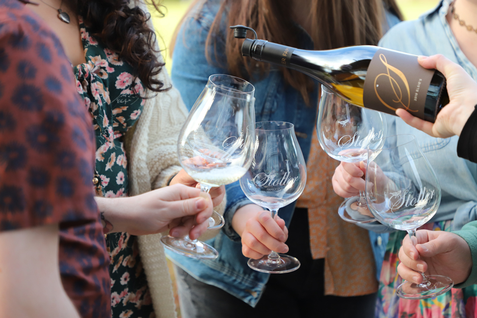 tasting room staff member pours Chardonnay into wine glasses held by four individuals