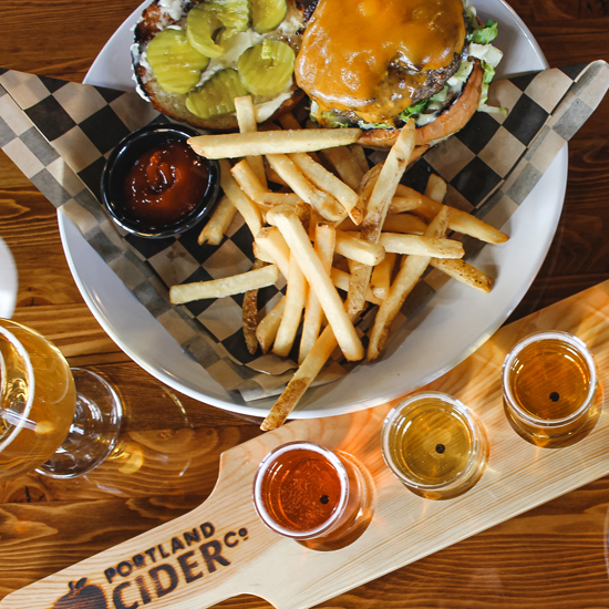 overhead shot of plated cheeseburger and fries next to stemmed glass of cider and taster tray of three ciders