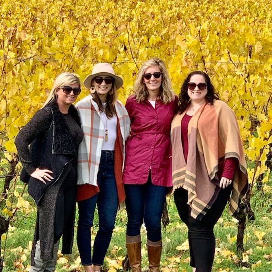 four people stand close together and smile at camera in front of leafy wine vines