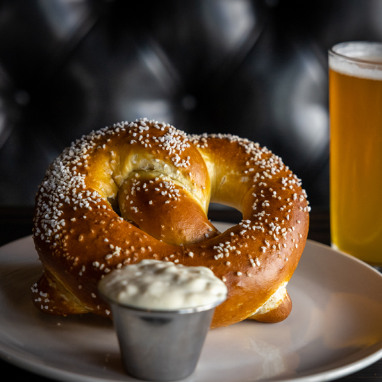 soft pretzel on plate with small container of dipping sauce and pint of beer next to plate