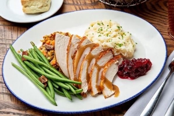 Sliced roasted turkey with gravy, mashed potatoes garnished with herbs, green beans, stuffing, an cranberry sauce on a white plate.