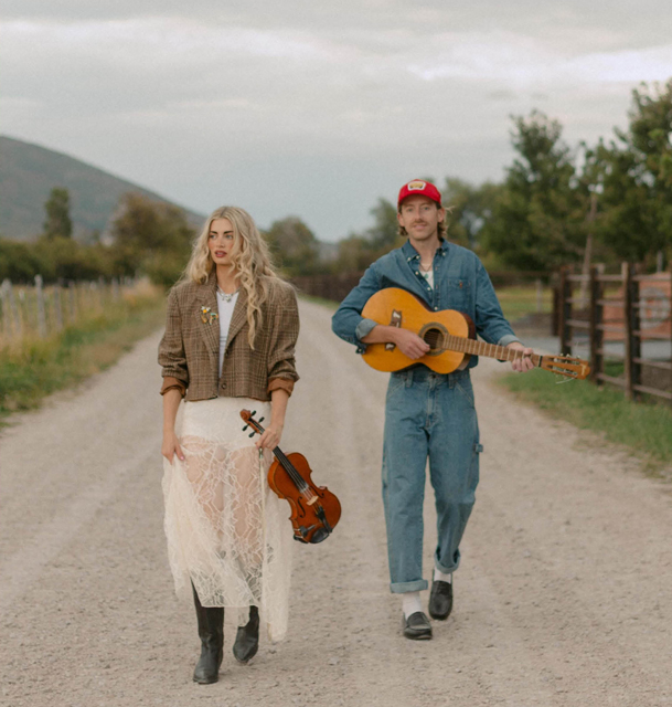 Two musicians walking down a rural dirt road with trees and mountains in the background.