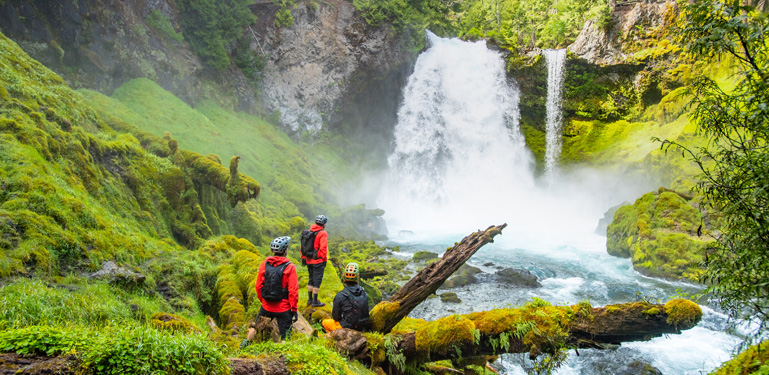 three people wearing bicycle helmets look upon a waterfall
