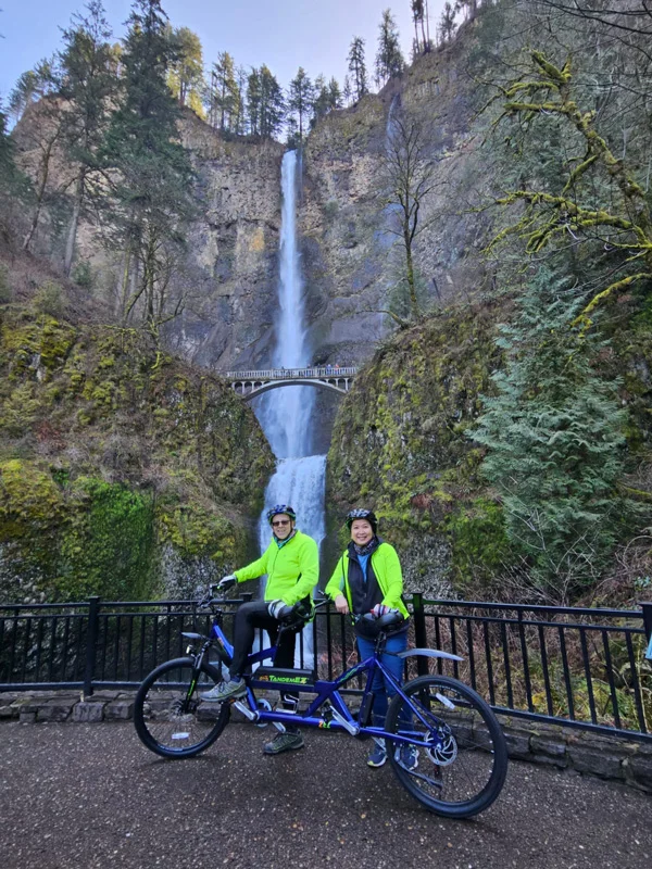 Two cyclist pose with their tandem e-bike in front of a tall waterfall.