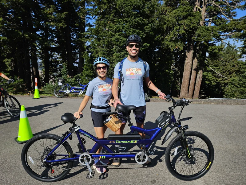 Two people wearing bicycle helmets smile while holding a tandem e-bike.
