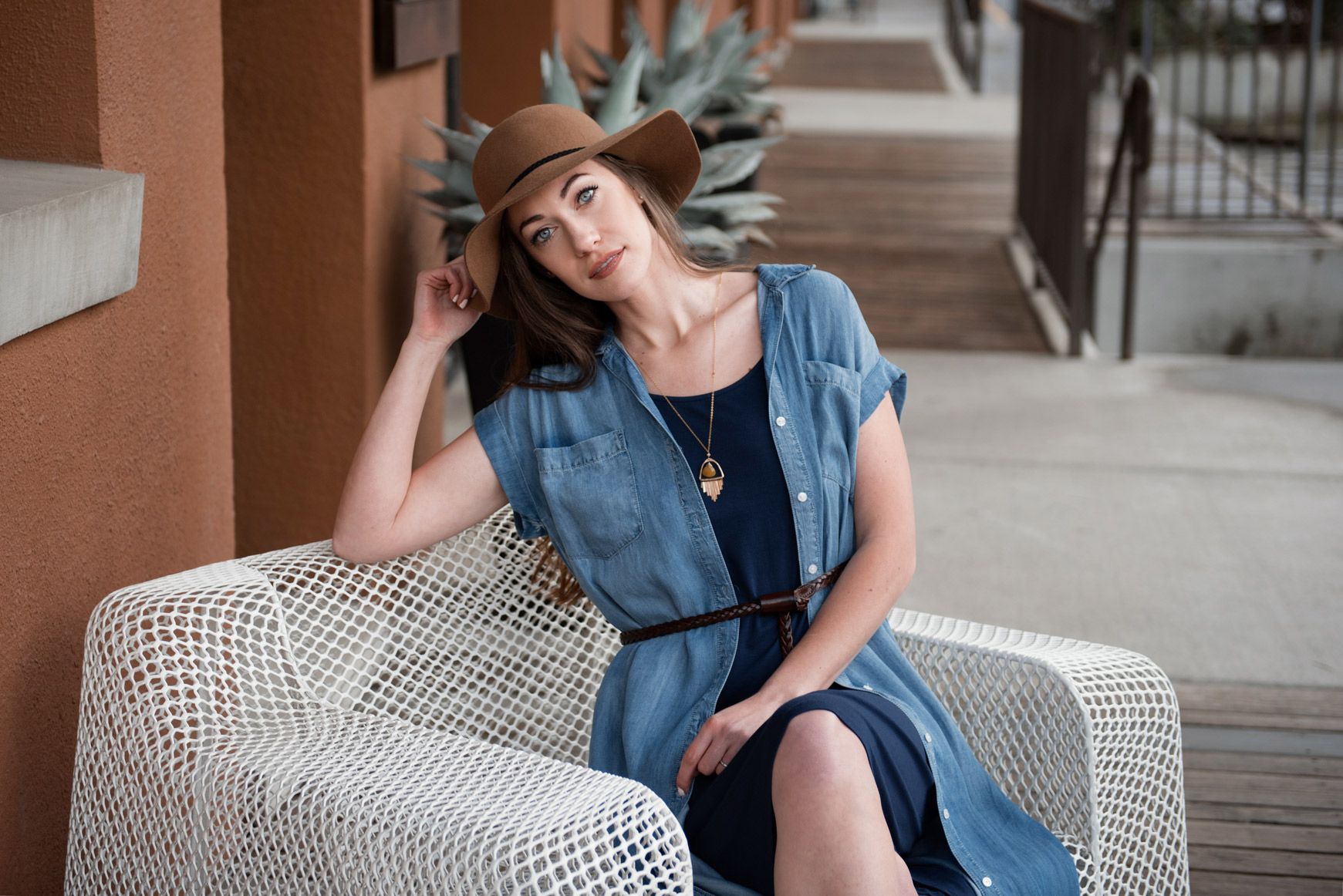 Woman in denim dress and hat sitting on a white chair.