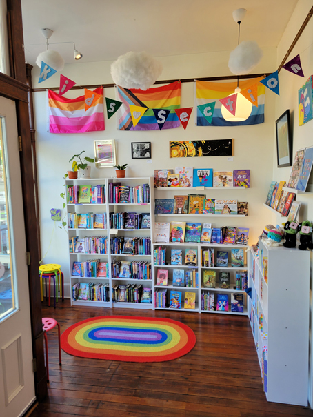 A cozy reading corner in a bookstore or library decorated with LGBTQ+ pride flags and a colorful "Kid's Section" banner.