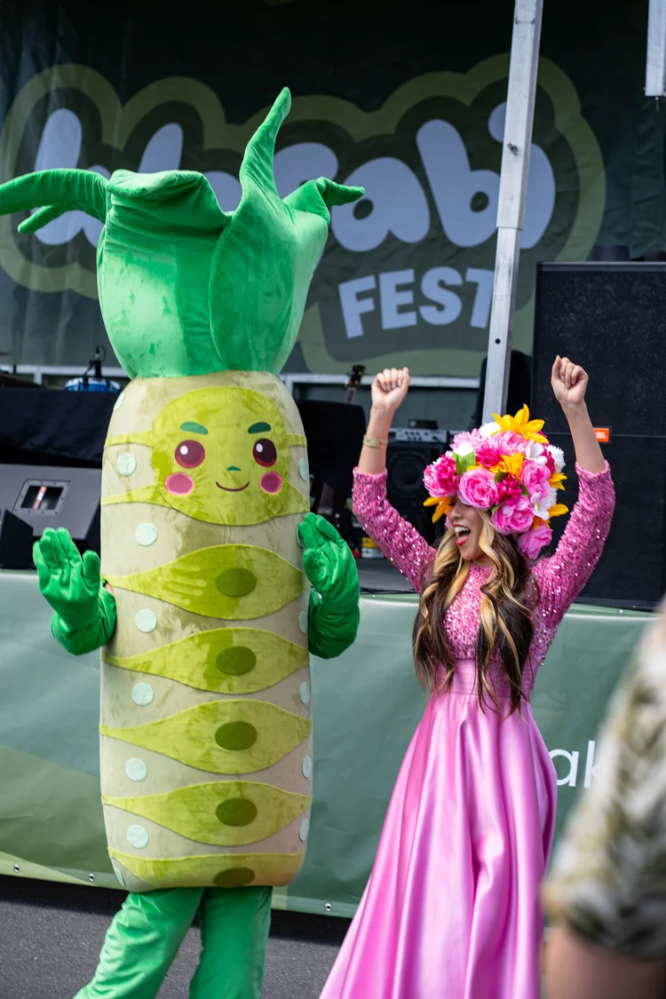 Person wearing a wasabi character mascot suit stands next to a woman wearing a pink sparkly dress and flower hat with her hands in the air.