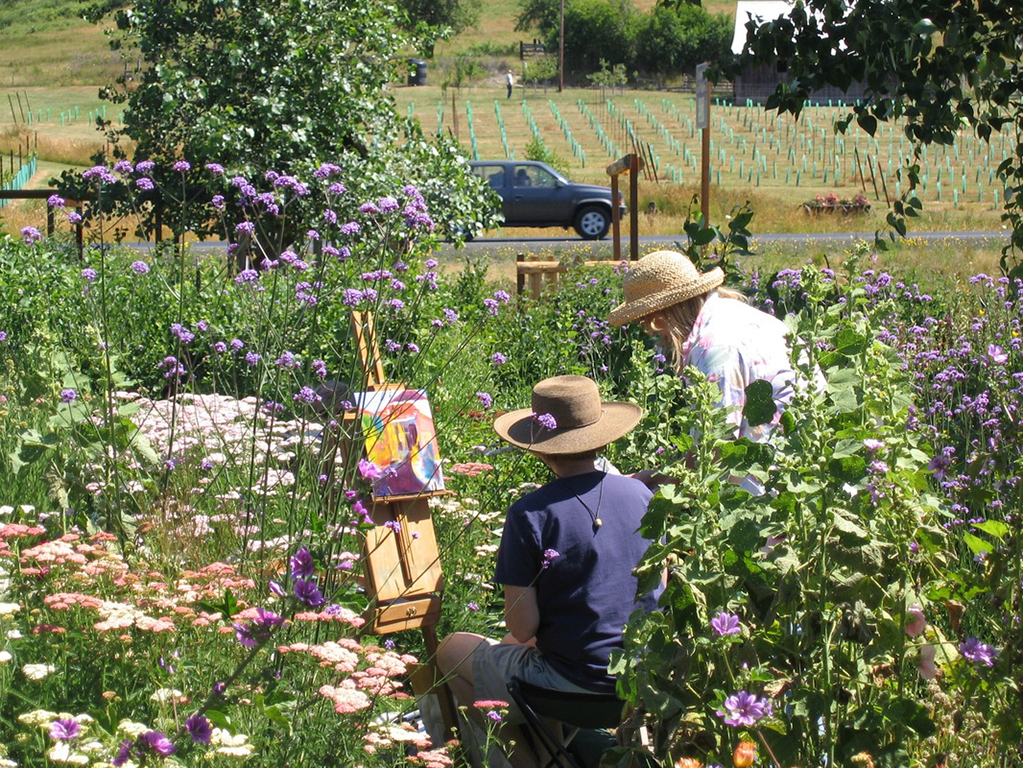 two people wearing sun hats outdoors in a patch of flowers, one sitting near easel, the other standing