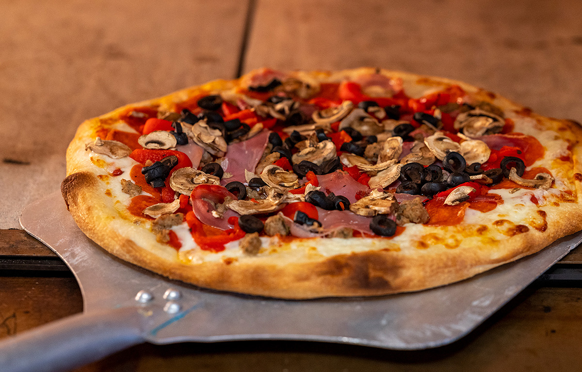 Specialty pizza from Timbers Cafe topped with pepperoni, ham, mushrooms, black olives, sausage and red peppers, baked on a golden crust and served on a pizza peel.