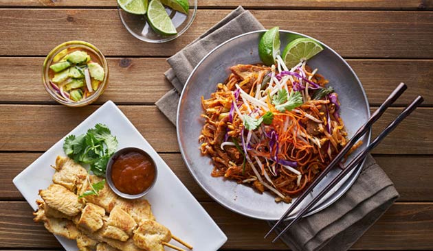 Grilled chicken skewers, stir-fried noodles, and a lime garnish on a wooden table.