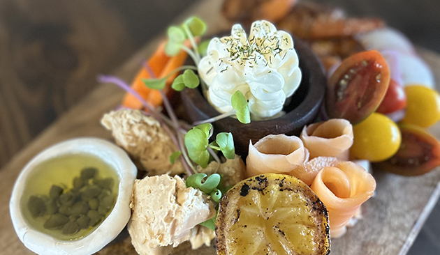 Charcuterie-style appetizer from Wildflour Public House featuring smoked salmon, cured meat, deviled egg, tomatoes, capers, grilled lemon, vegetables and fresh herbs on a wooden board.