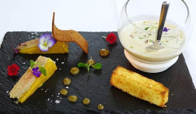 Elegant dish with yellow dessert, soup, and garnishes on black slate.