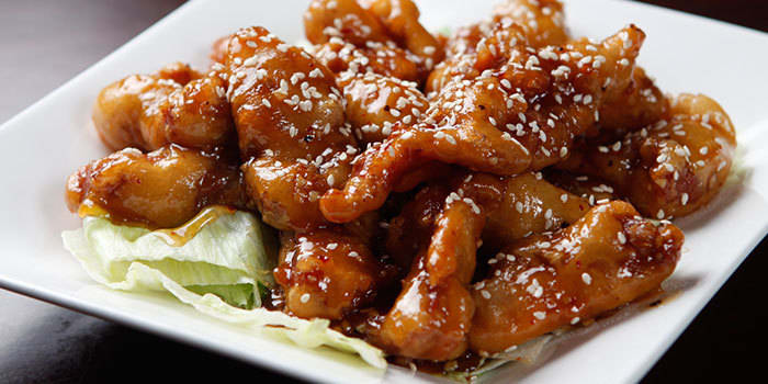 Sesame chicken from Tai's Dynasty served on crisp lettuce with a glossy sauce and sesame seeds on a white square plate.