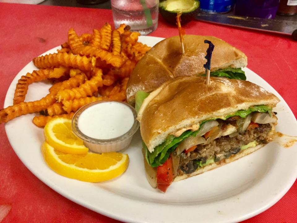Burger from The Tin Thistle topped with grilled onions, lettuce, tomato and cheese, served with waffle fries, dipping sauce and orange slices on a white plate.