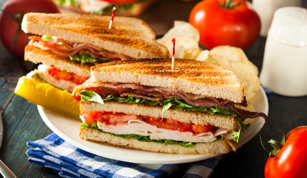 Blue Heron Bistro serves toasted club sandwiches layered with sliced turkey, bacon, lettuce and tomato, accompanied by potato chips on a white plate.