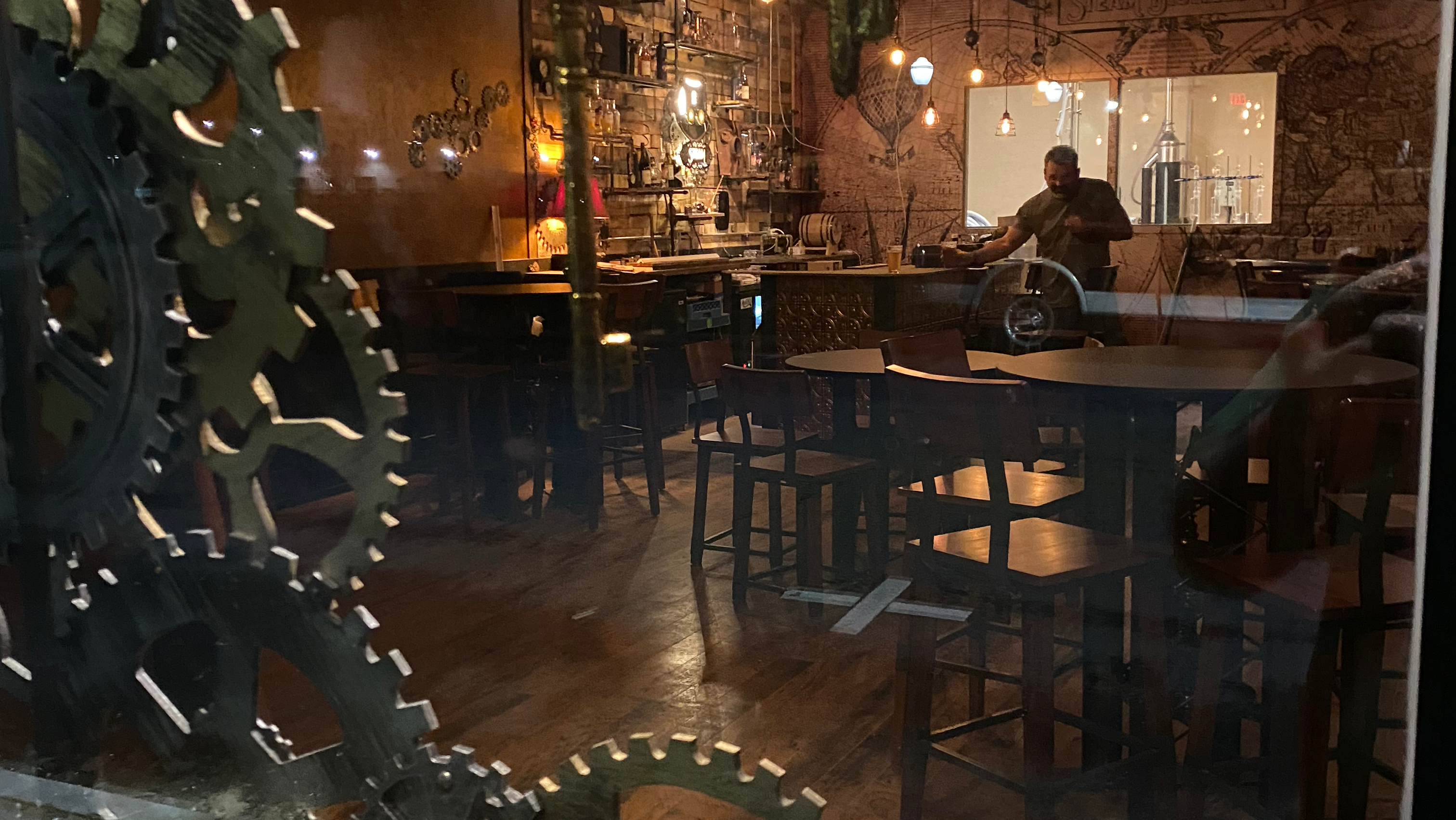 A dimly lit distillery tasting room interior featuring wooden tables and chairs, with large decorative gears in the foreground.