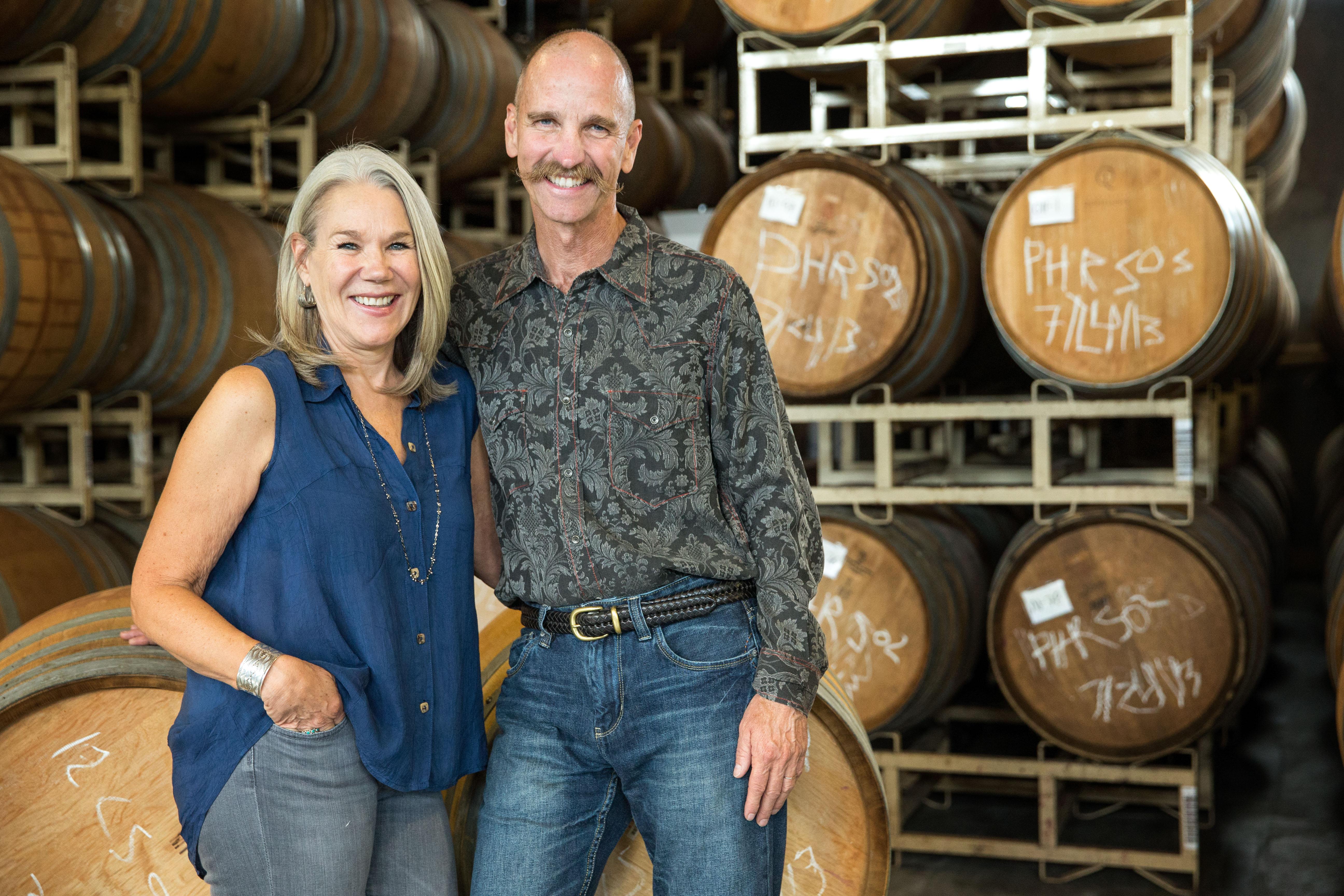 two people smile while posting in front of barrels in a wine cellar