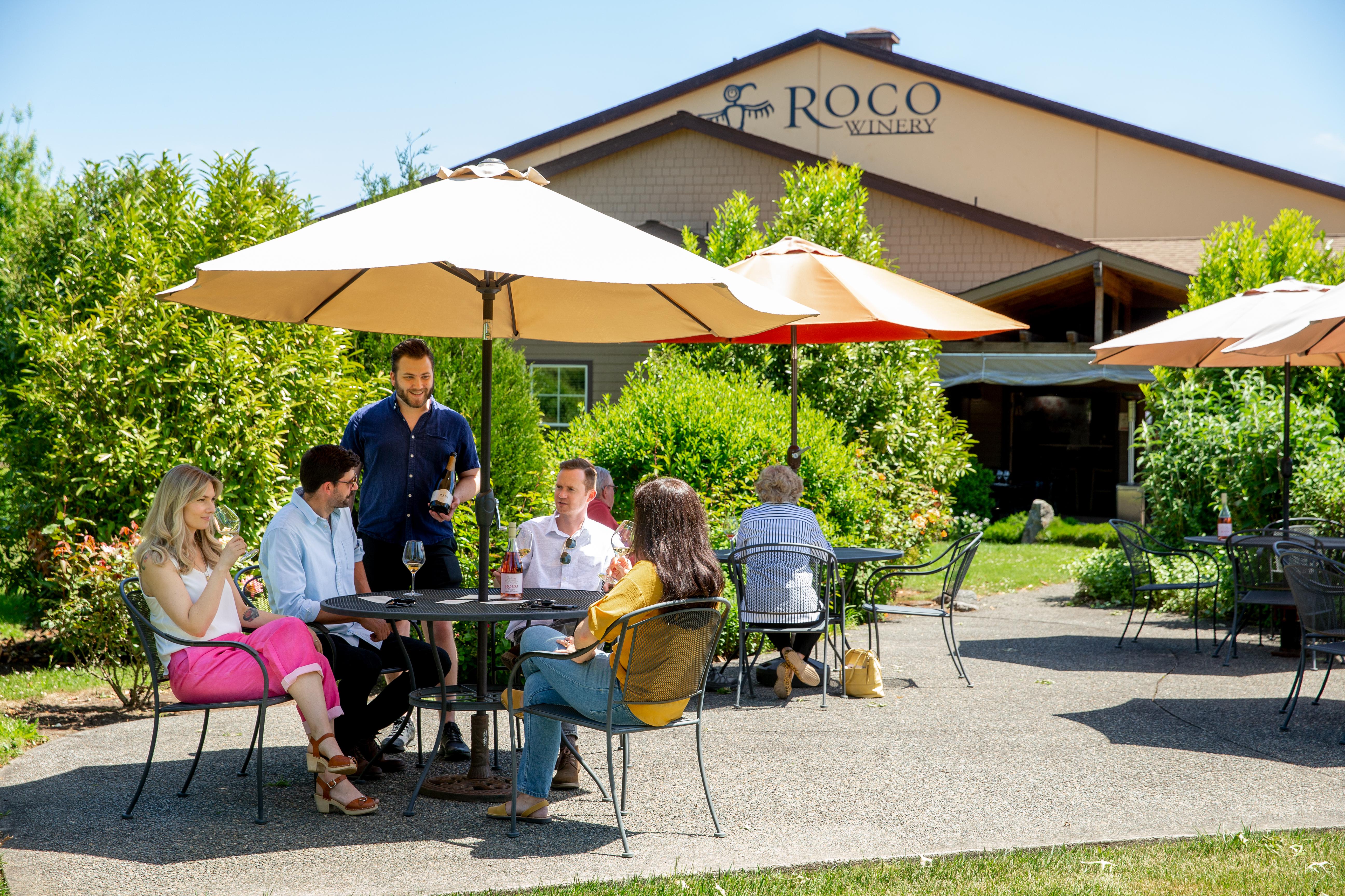 people seated at patio table with umbrella outside of building with sign for ROCO winery