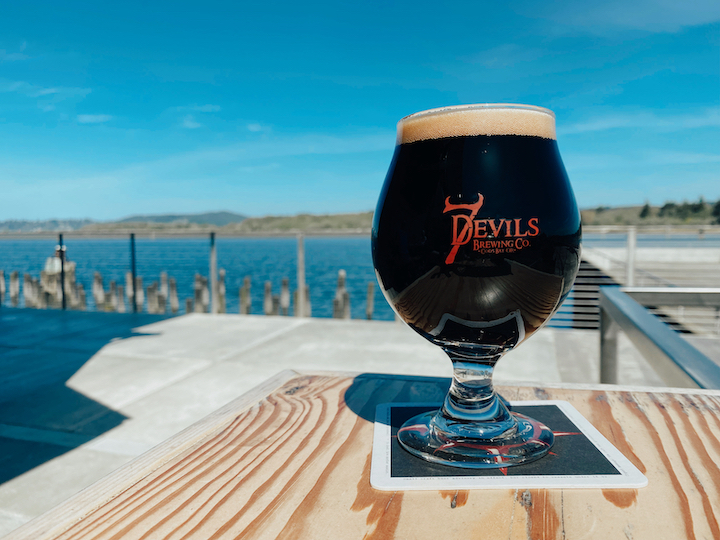 7 Devils Waterfront Alehouse features a dark craft beer in a branded glass on an outdoor table overlooking Coos Bay, with waterfront views and blue sky in the background.