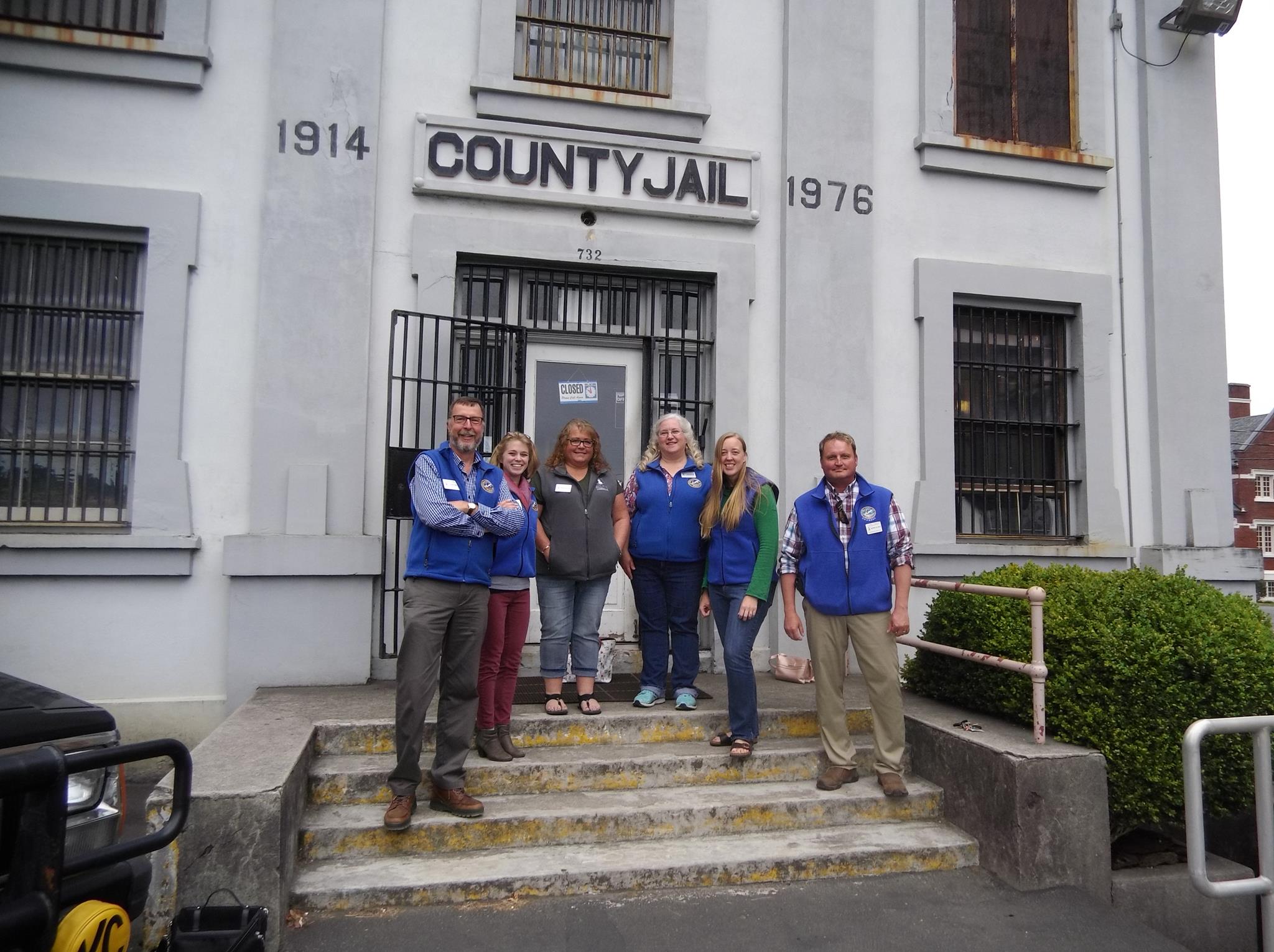 six people pose for photo in front of a historic county jail building that is now a museum