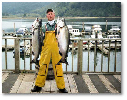 person standing on dock wearing waders and holding a large fish in each hand