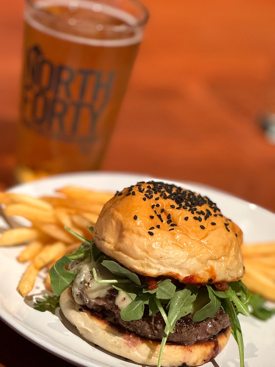 Burger and fries served at North Forty Brewery, topped with arugula, melted cheese and bacon on a seeded bun, with a pint of beer blurred in the background.