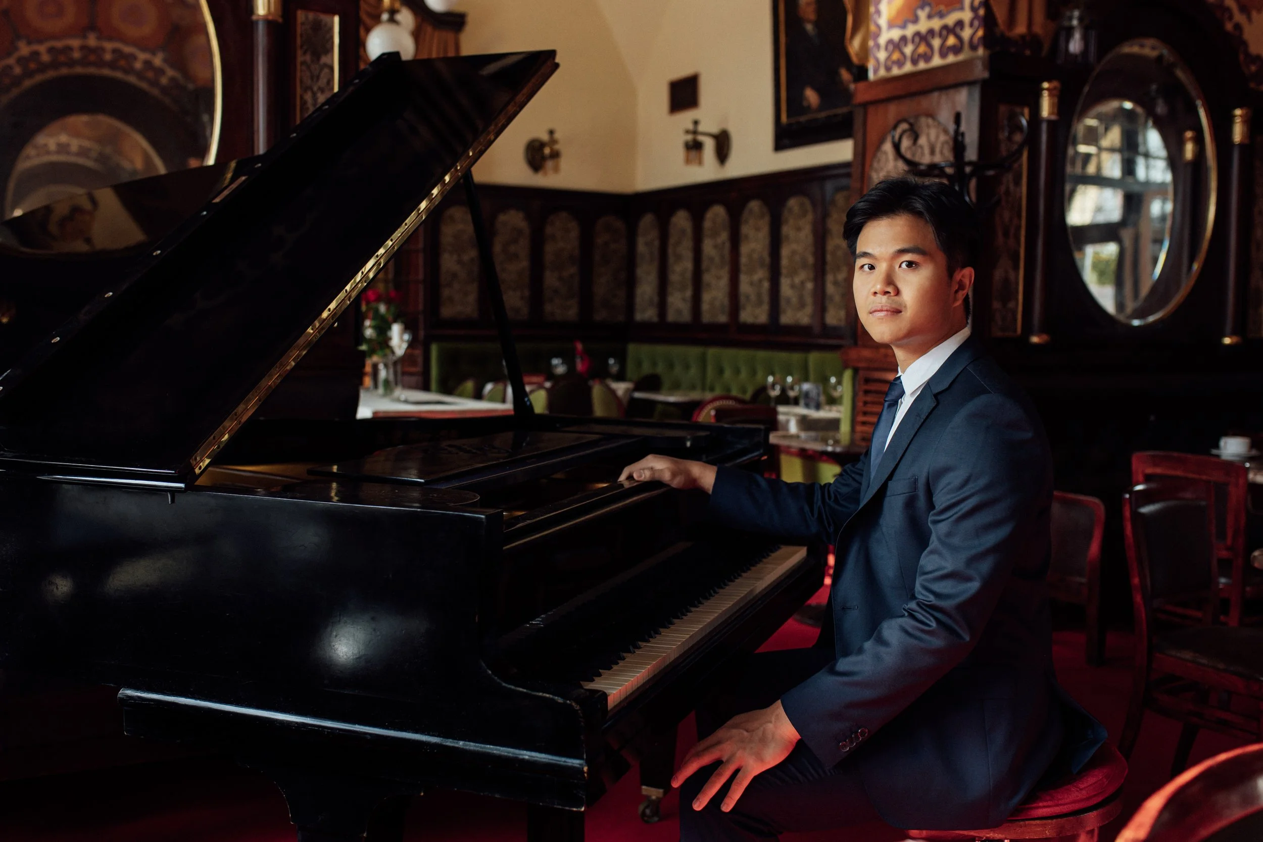 person wearing a dark suit and matching time with a white collared shirt sitting on a stool in front of a piano