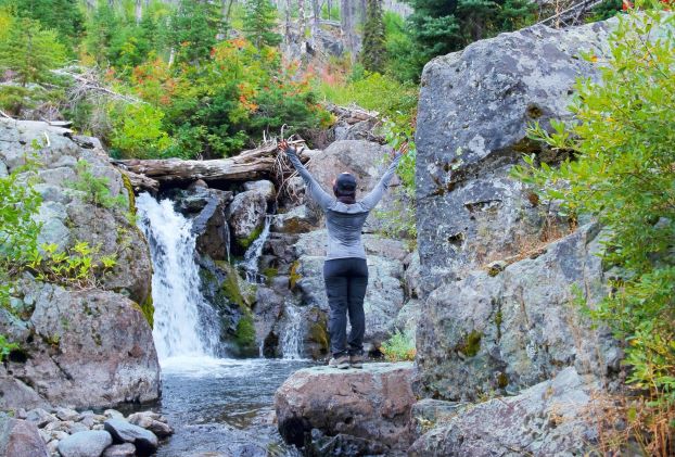 woman standing on rock with facing a waterfall with her arms up in the air