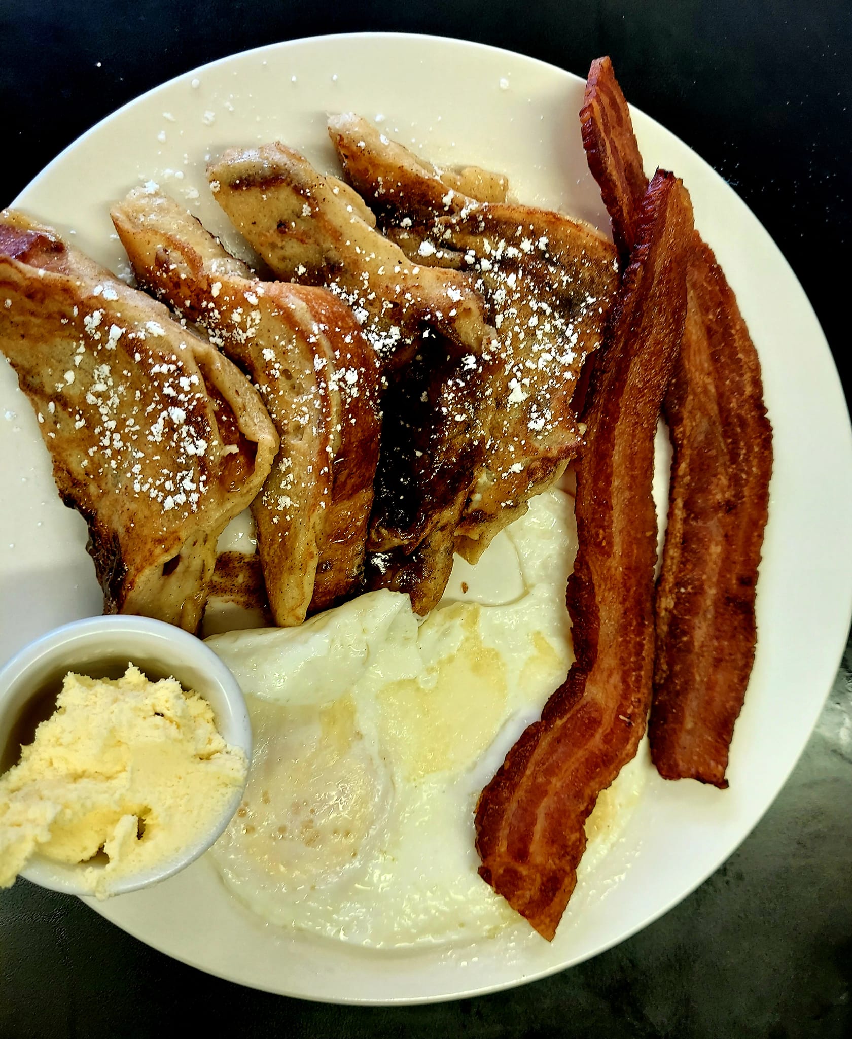 french toast dusted with powdered sugar, two eggs over medium and two strips of bacon on a white plate