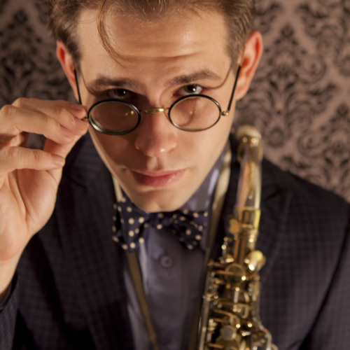 man holding a saxaphone in one hand and looking over his glasses, aaron johnson press photo