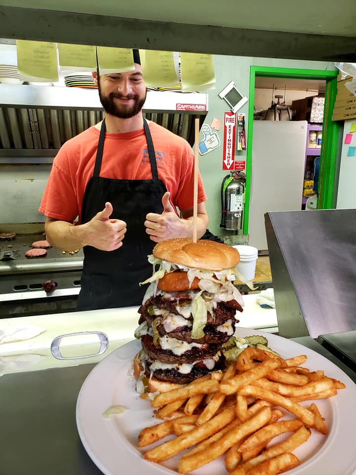 chef in kitchen giving two thumbs up at the plated 5 patty burger and fries sitting in the window