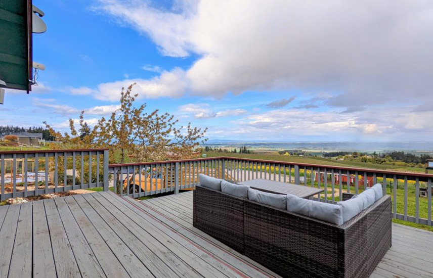 An outdoor sofa and table is on a wood deck with rails.  Beautiful views and a sky with fluffy clouds are in the distance.