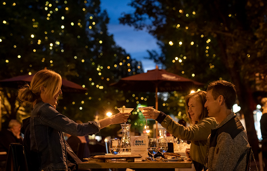 Two women and a man cheers at a table under the twinkle lights on 3rd Street