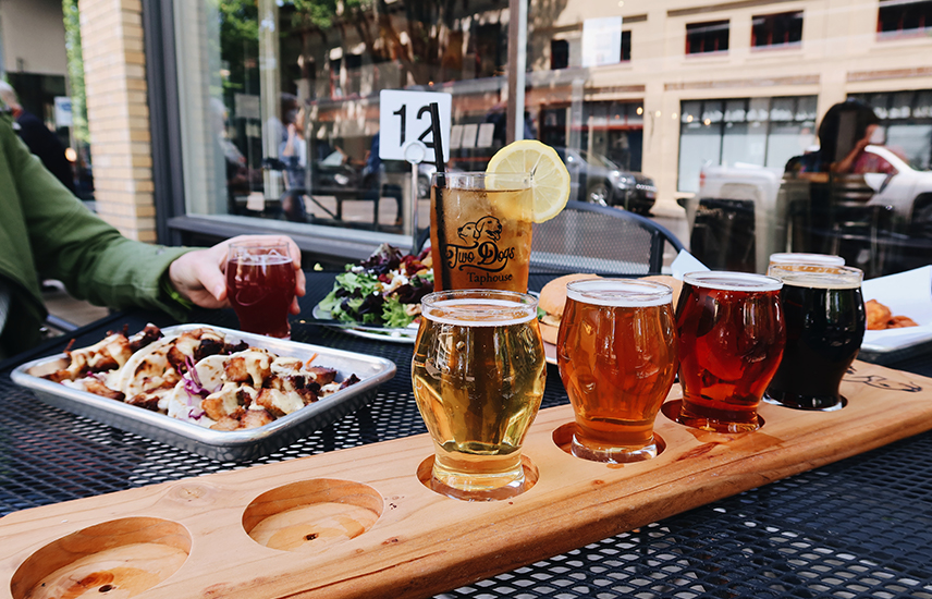 A flight of beer is lined up on an outdoor table.  Plates of food are in the background.