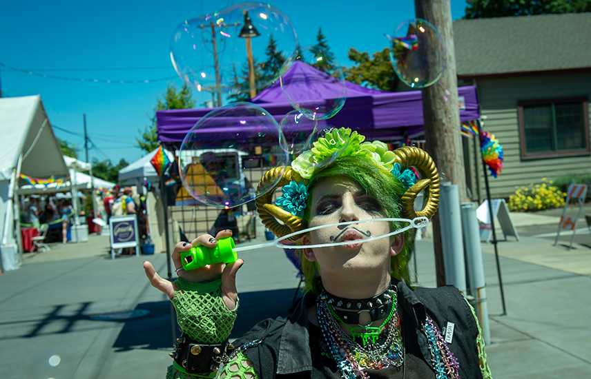 A person in a green costume blows bubbles.  Tents with vendor wares are in the background.
