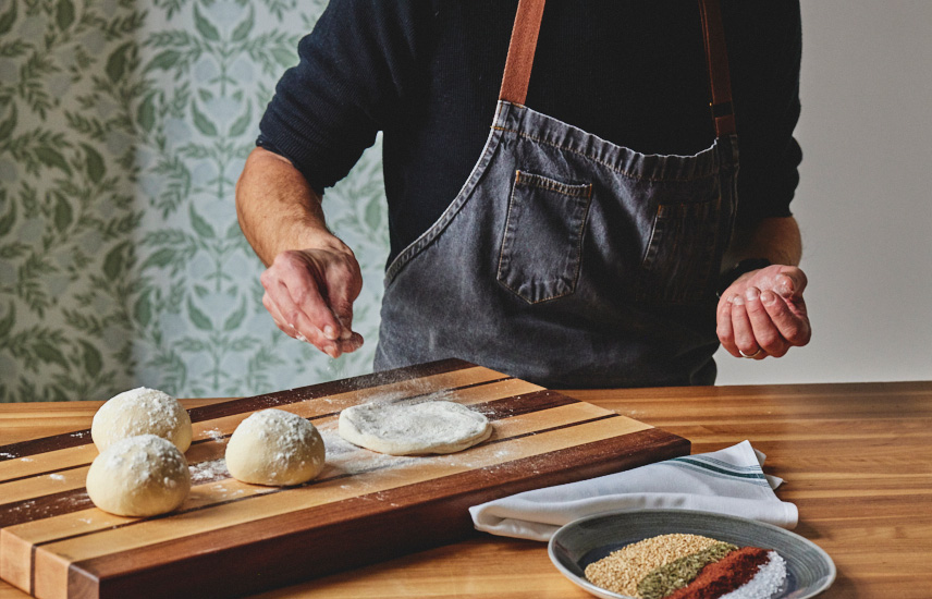 A man sprinkles flour onto dough balls and a disc of dough.  A plate full of spices and a cloth napkin are next to them.