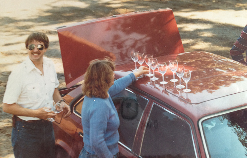 A retro photo of a man and woman lining up wine glasses on top of the roof of a car.