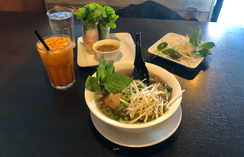 A bowl of beef pho is topped with bean sprouts and basil.  It is accompanies by shrimp salad rolls and Thai iced tea.