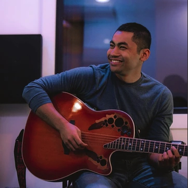 A Man sits, playing the guitar and smiles.
