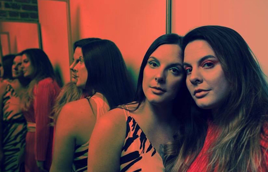 Two women look at the camera.  The back of their reflection is behind them.
