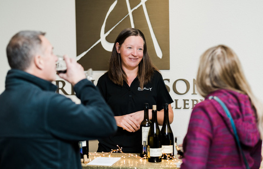 A woman smiles.  She stands in front of a table with bottles of wine.  Two guests sample the wine.