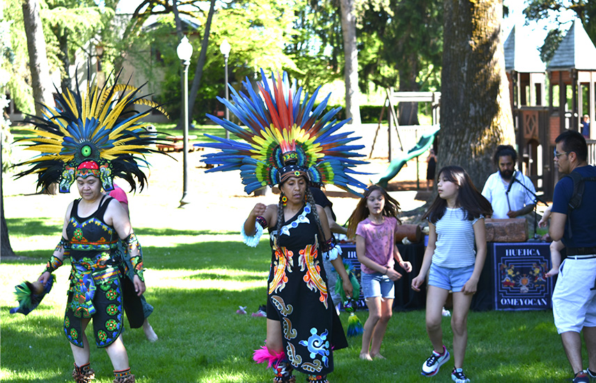 Two women with feathered headdresses demonstrate a dance in the park.