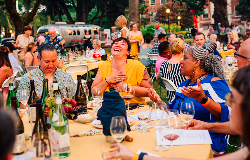 Several people of color sit at an outdoor table, laughing and drinking wine.