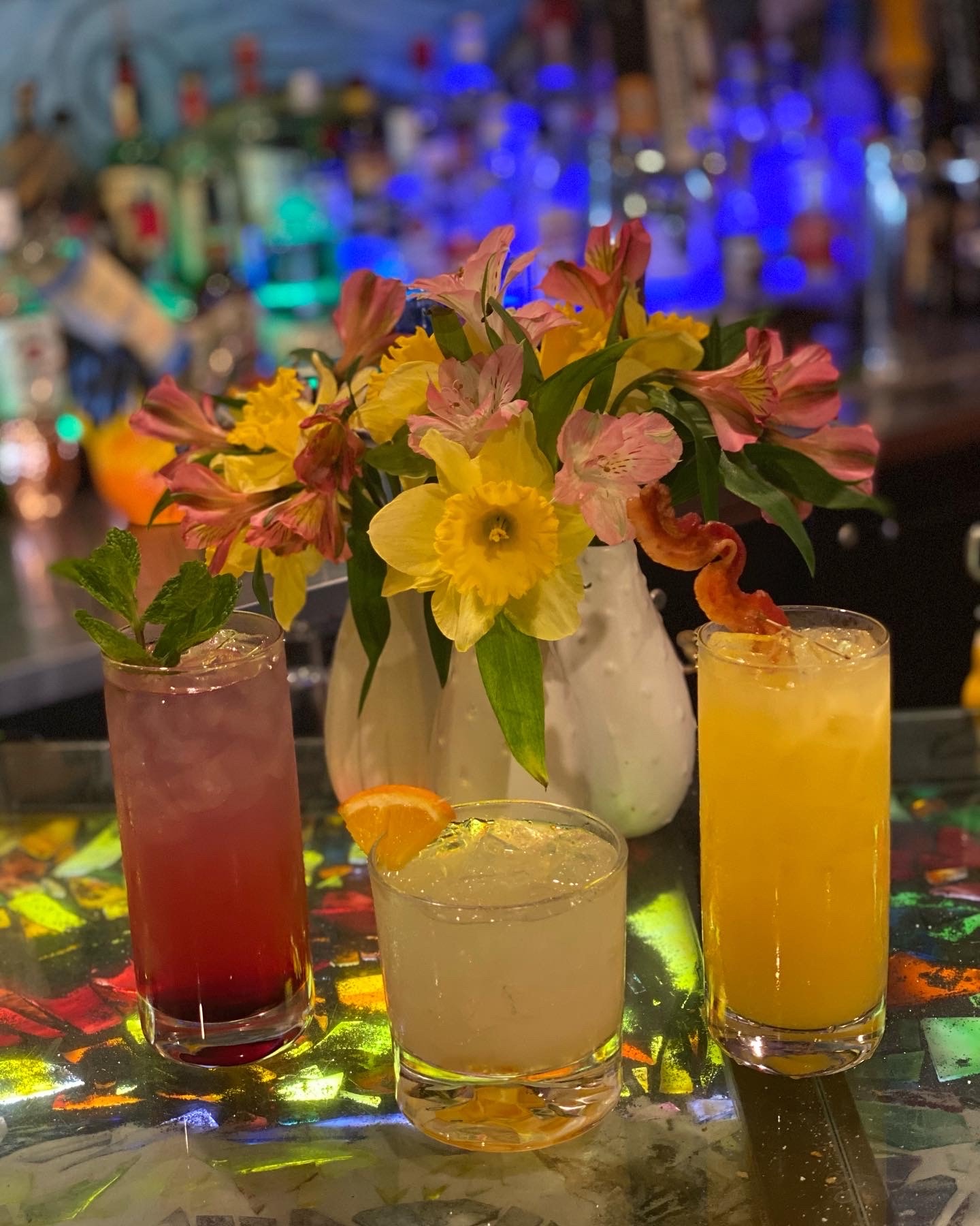 three glasses of colorful spirits displayed on bar top in front of vase of flowers