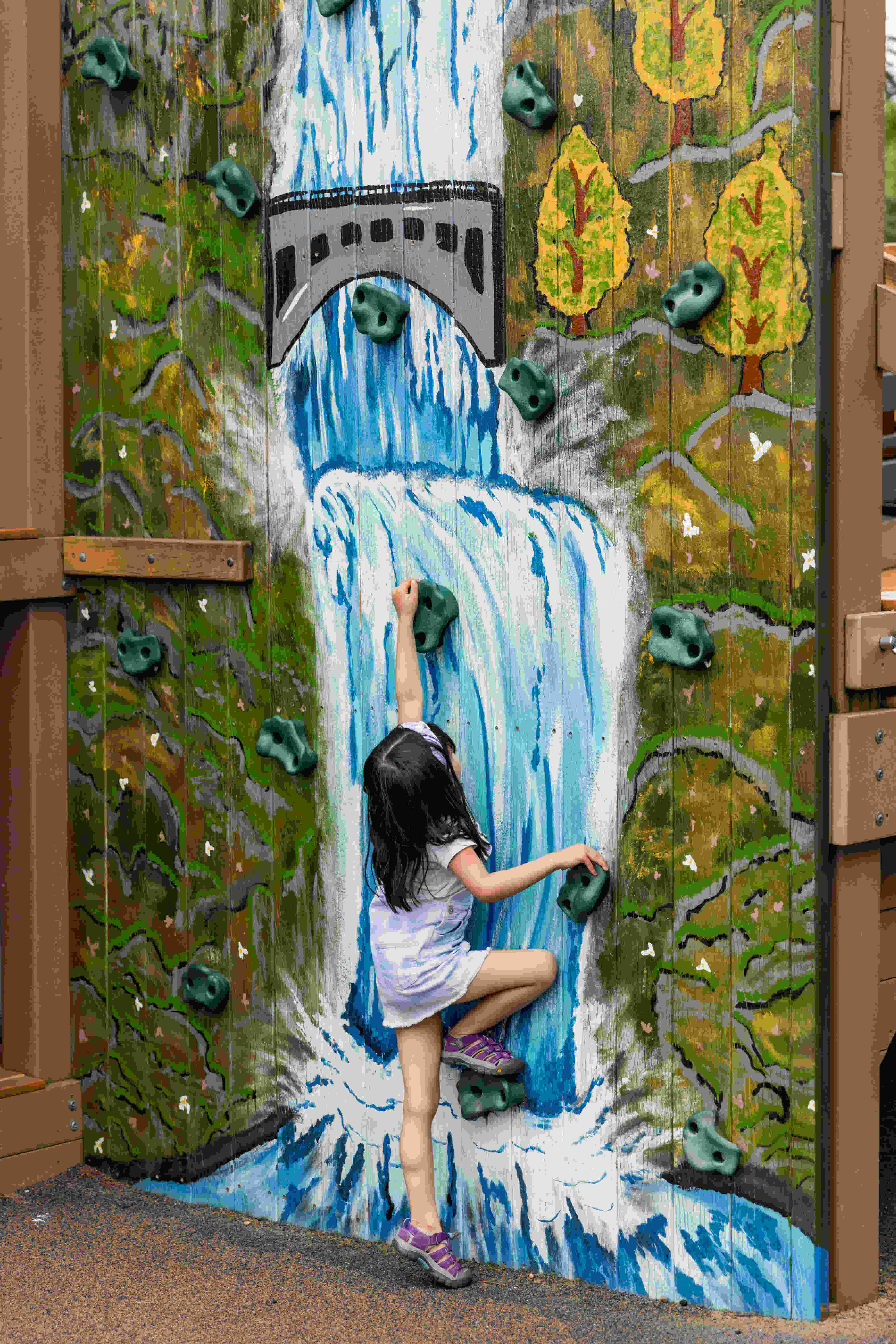 Young girl climbs up outdoor rock wall with Multnomah Falls painted on it.