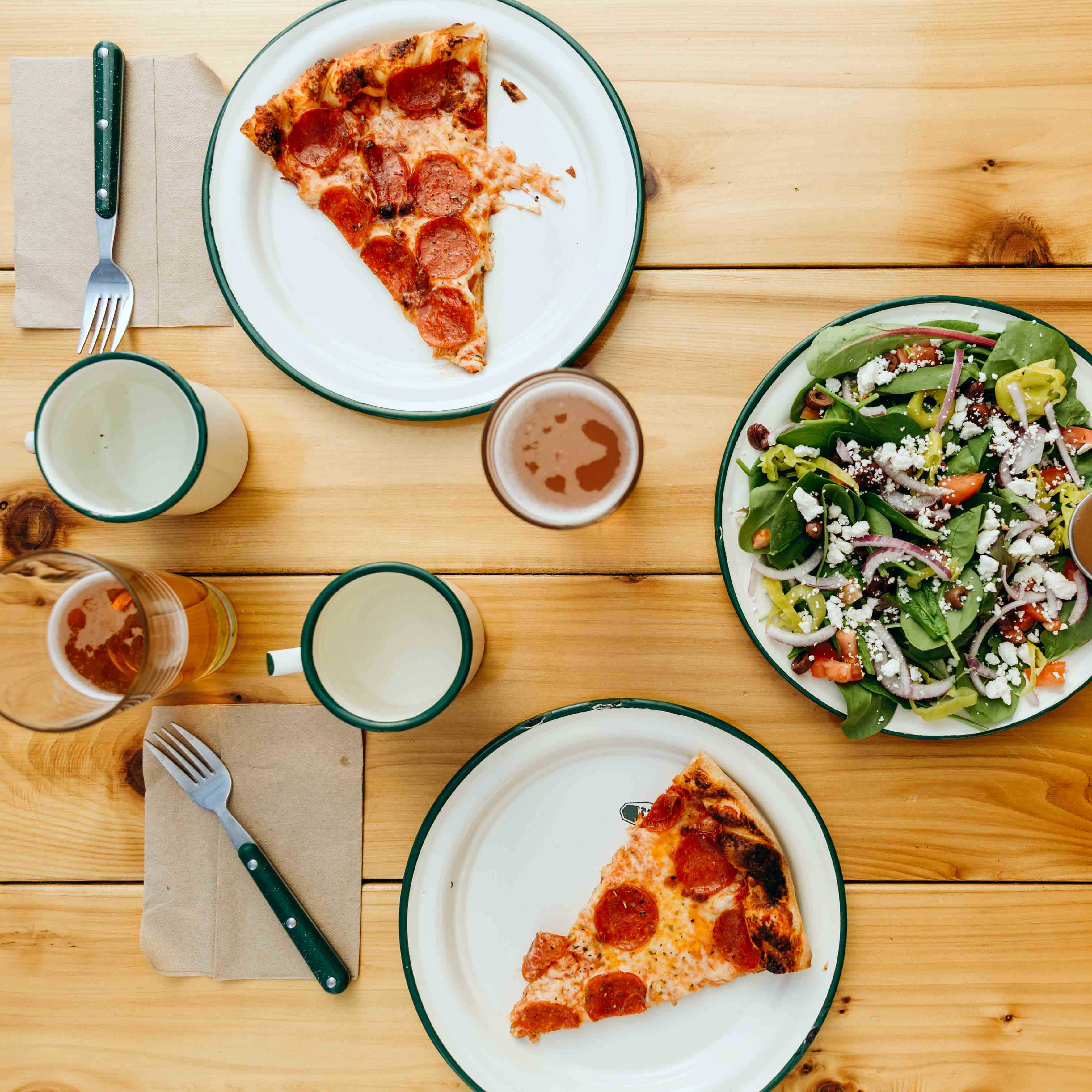 table set with enamelware plates and mugs holding slices of pepperoni pizza and fresh salad, accompanied by beer.
