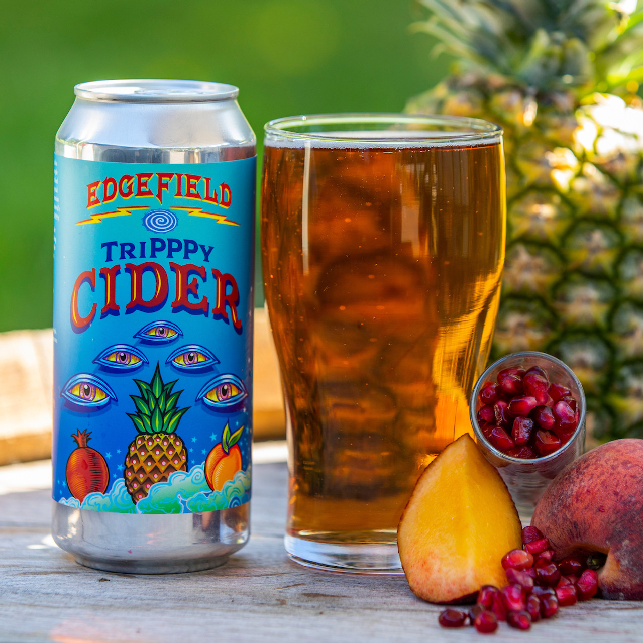 Can of Edgefield Tripppy Cider next to draft pour of it and a pineapple, pomegranate, and peach